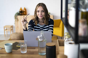 Smiling woman holding eyeglasses in front of laptop at kitchen - GIOF14022