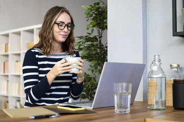 Lächelnde Frau mit Kaffee vor einem Laptop an der Küchentheke - GIOF14023