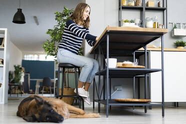 Lächelnde Frau auf einem Hocker neben einem Hund, der sich in der Küche entspannt - GIOF14029