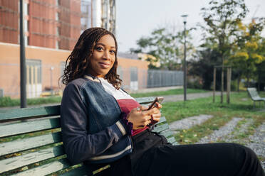 Lächelnde Frau mit Mobiltelefon auf einer Bank sitzend - MEUF04661