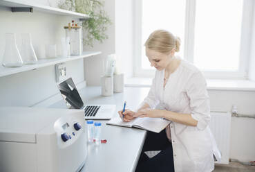 Blonde Wissenschaftlerin mit Laptop, die am Schreibtisch in einen Notizblock schreibt - AHSF02801