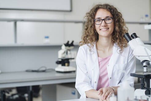 Lächelnde junge Wissenschaftlerin mit Mikroskop am Labortisch - AHSF02841