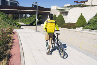 Delivery man with prosthetic leg wheeling bicycle on footpath - JCCMF04334