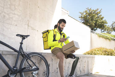 Smiling disabled delivery man scanning box with mobile phone by bicycle - JCCMF04343