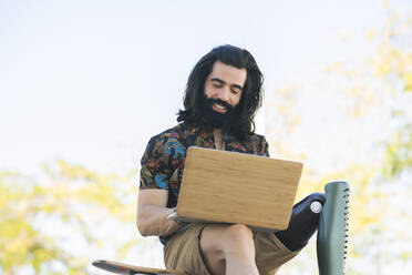 Smiling disabled man with long hair using laptop in public park - JCCMF04354