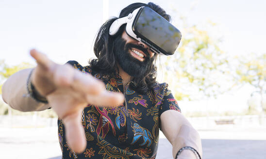 Lächelnder Mann mit virtuellem Headset gestikuliert in einem öffentlichen Park - JCCMF04356