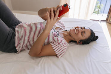 Smiling woman using mobile phone on bed at home - JRVF02019