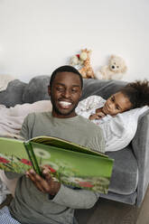 Lächelnder Mann, der vor seiner Tochter zu Hause ein Buch liest - VYF00602