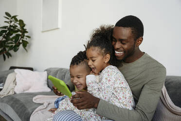 Cheerful son and daughter watching digital tablet with father on sofa - VYF00616