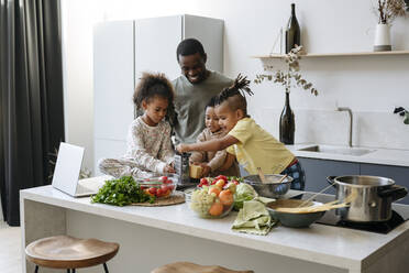 Smiling father looking at sons and daughter grating cheese in kitchen - VYF00628