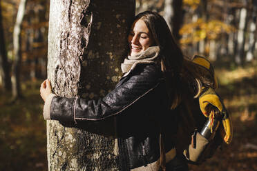 Happy young woman hugging tree in autumn forest - MCVF00928