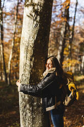 Smiling young woman hugging tree in autumn forest - MCVF00929