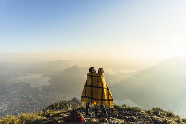 Women wrapped in blanket looking over shoulder on mountain peak at Lecco, Italy - MCVF00938