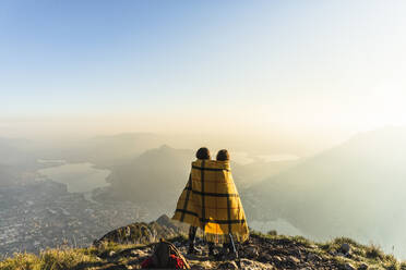 Friends wrapped in blanket on mountain peak at Lecco, Italy - MCVF00939