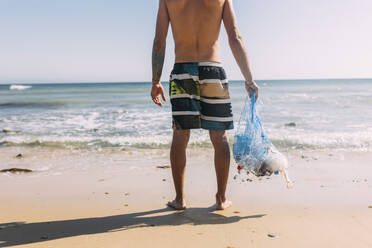 Shirtless male volunteer holding mesh bag at beach - EGHF00244