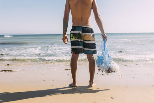 Männlicher Freiwilliger ohne Hemd, der eine Netztasche am Strand hält - EGHF00244