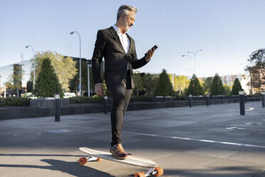 Geschäftsmann steht auf einem Skateboard und benutzt ein Smartphone in der Stadt - JCCMF04382