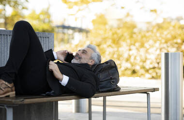 Businessman lying on bench talking on smart phone - JCCMF04393