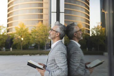 Businessman holding book leaning on glass wall - JCCMF04422