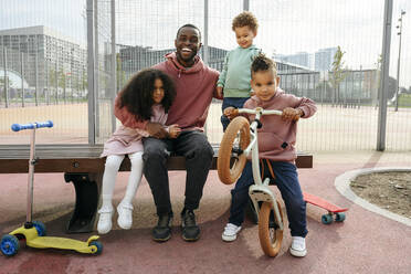 Glücklicher Vater sitzt mit Arm um Kinder auf dem Sportplatz - VYF00691
