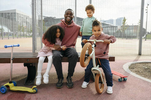 Happy father sitting with arm around children at sports field - VYF00691