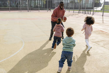 Fröhlicher Vater spielt Basketball mit Kindern auf einem Sportplatz - VYF00692