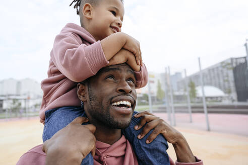 Lächelnder Vater schaut auf seinen Sohn, der auf den Schultern im Sportplatz sitzt - VYF00707