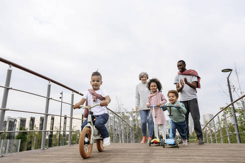 Father cheering for children riding push scooter and bicycle on bridge - VYF00736