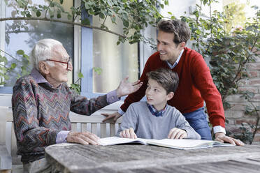 Smiling man looking at father explaining to grandson studying in backyard - AANF00177