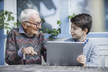 Smiling boy with laptop talking with grandfather at table - AANF00182