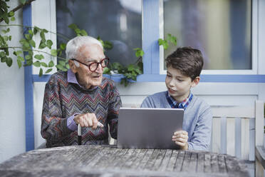 Senior man talking to grandson sitting with tablet PC at table - AANF00184