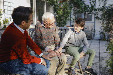 Caring boy and father touching hands of grandfather in backyard - AANF00199