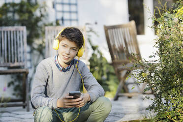 Lächelnder Junge mit Mobiltelefon hört Musik über Kopfhörer bei einer Pflanze - AANF00204