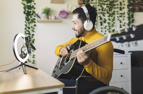 Smiling male influencer playing guitar on wheelchair - JCCMF04440