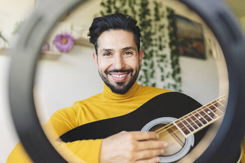 Smiling male influencer playing guitar seen through ring light - JCCMF04442