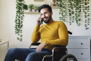 Lächelnder Mann mit Behinderung, der zu Hause mit seinem Smartphone telefoniert - JCCMF04445