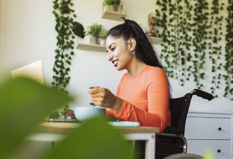 Lächelnde behinderte Frau mit Essen und Laptop zu Hause - JCCMF04446