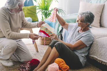 Senior woman showing knitted wool to man in living room - SIPF02680