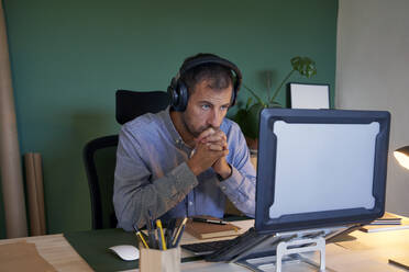Nachdenklicher Geschäftsmann mit Kopfhörern, der im Heimbüro auf seinen Laptop schaut - VEGF05132