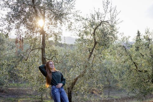 Lächelnde Frau lehnt sich bei Sonnenuntergang an einen Olivenbaum - EIF02351