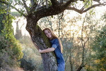 Smiling young woman hugging tree at countryside - EIF02358