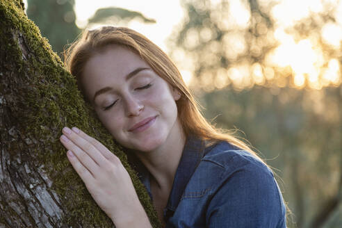 Frau mit geschlossenen Augen umarmt Baum bei Sonnenuntergang - EIF02359