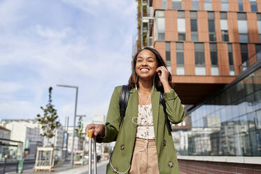 Smiling woman with luggage wearing headphones in city - KIJF04283