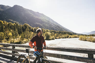 Man with mountain bike looking at river and mountains on bridge - UUF25026