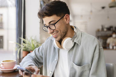 Lächelnder Geschäftsmann mit Brille, der in einem Café ein Mobiltelefon benutzt - JCCMF04464