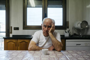 Sad man sitting at table in kitchen - JRFF05189