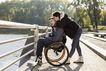 Girlfriend kissing boyfriend sitting in wheelchair at embankment - LLUF00291