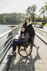 Woman embracing man sitting in wheelchair at embankment - LLUF00293