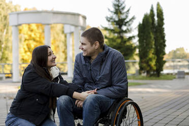 Freundin lächelt ihren Freund an, der im Rollstuhl im Park sitzt - LLUF00294