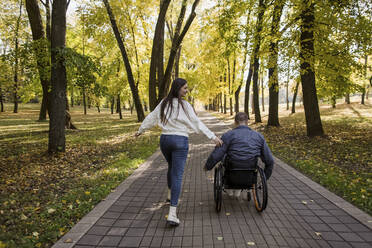 Verspielte Freundin läuft mit ihrem Freund im Rollstuhl sitzend im Park - LLUF00300